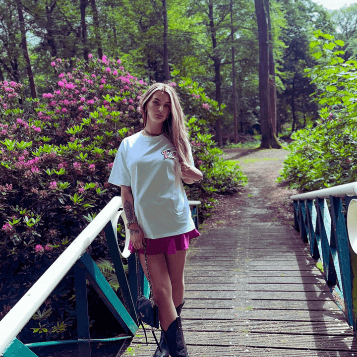 Woman wearing Daisy Flower T-Shirt with pink skirt, standing on a bridge in a lush green park with blooming flowers.