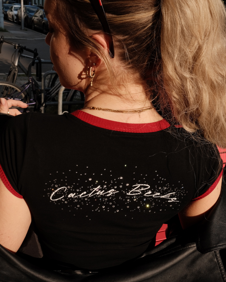 Woman wearing a black crop top with 'Cactus Becky' text in rhinestones and diamonds, standing outdoors.