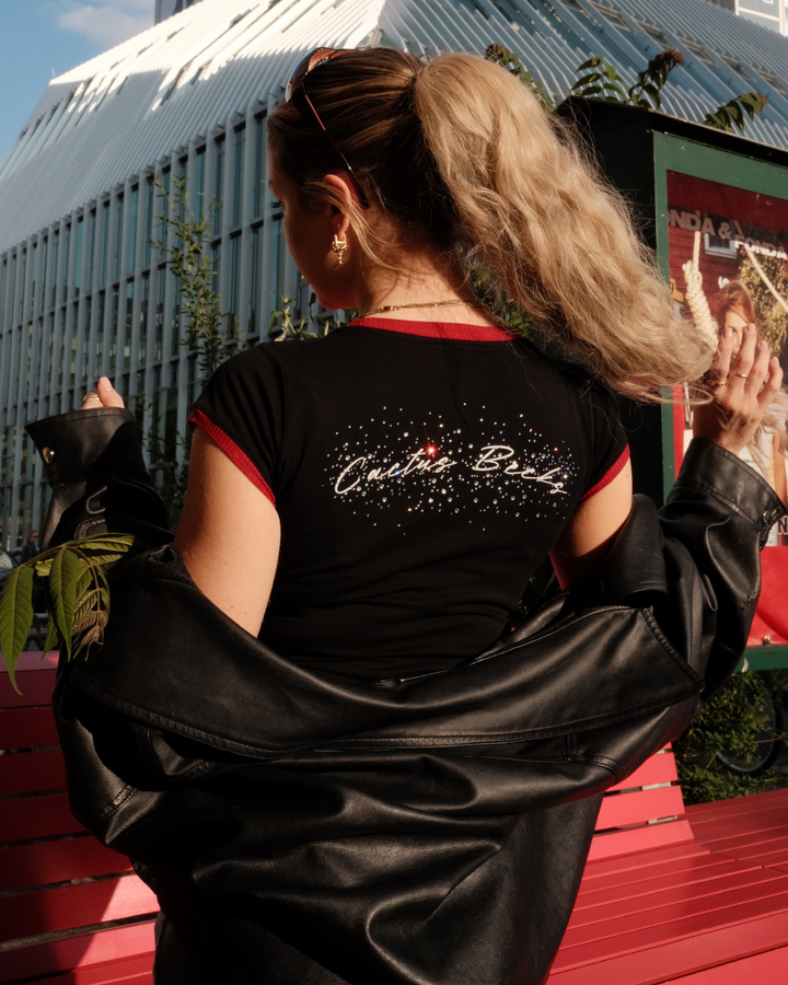Woman wearing a crop top and leather jacket with 'Cactus Becks' text covered in diamonds and rhinestones, standing outdoors.