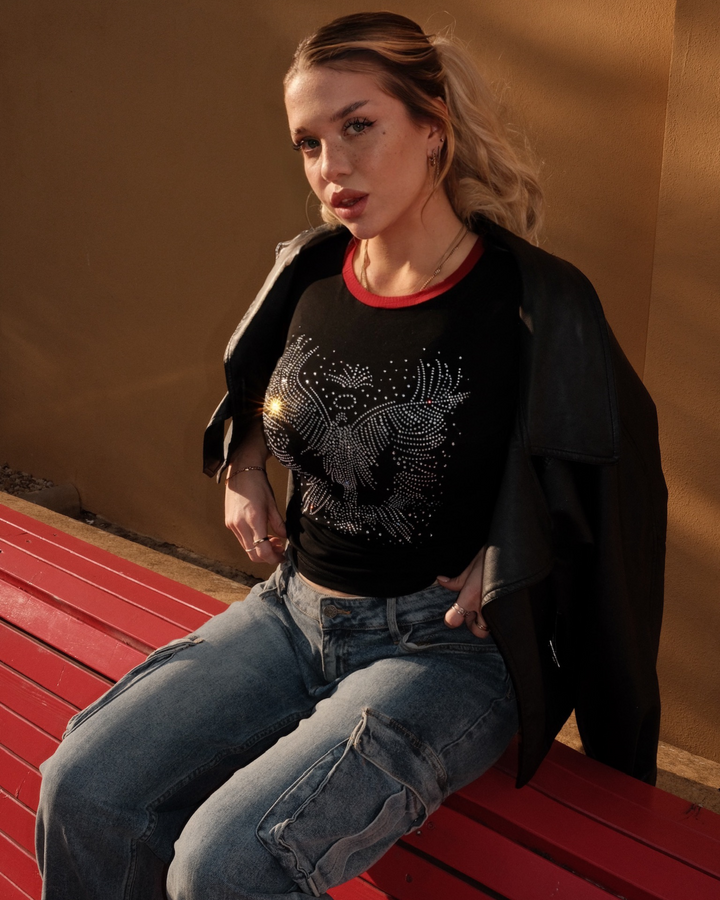 Woman sitting on a red bench wearing a black crop top with diamonds (rhinestones) and blue jeans.