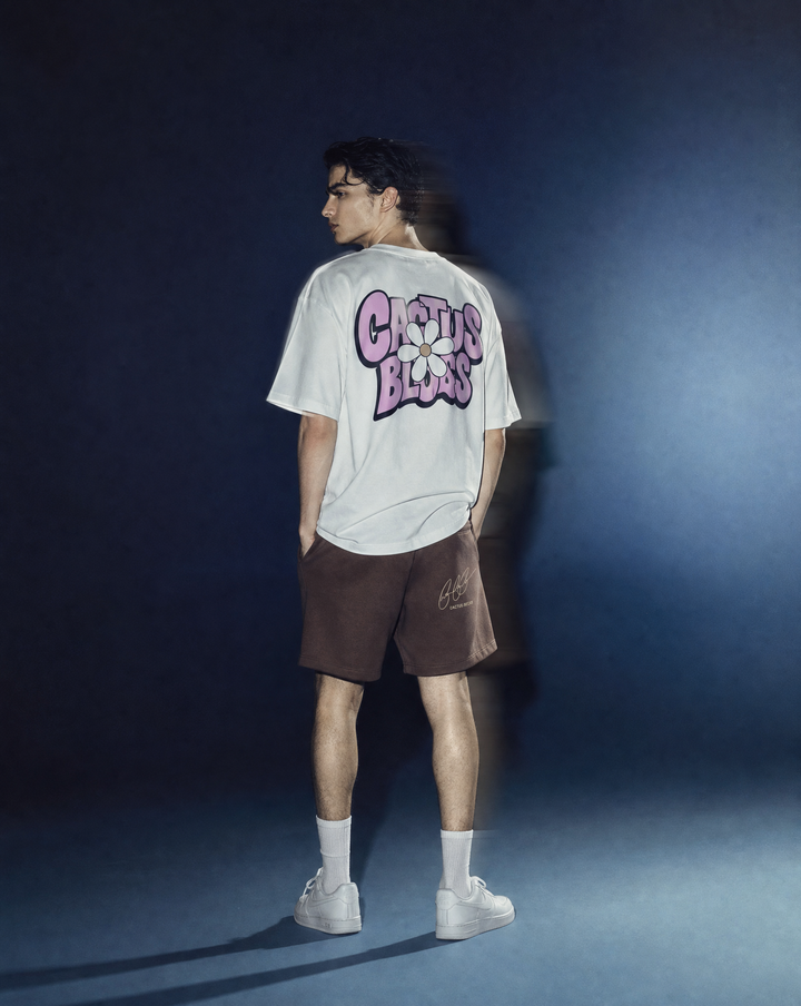 Person wearing a daisy flower t-shirt by Cactus Becks and a brown Cactus Becks shorts with a broken heart logo, against a blue background