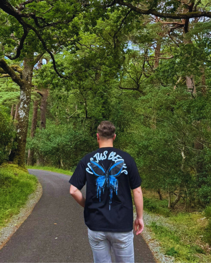 Person walking away on a path through a forest wearing a black t-shirt with a blue butterfly design.
