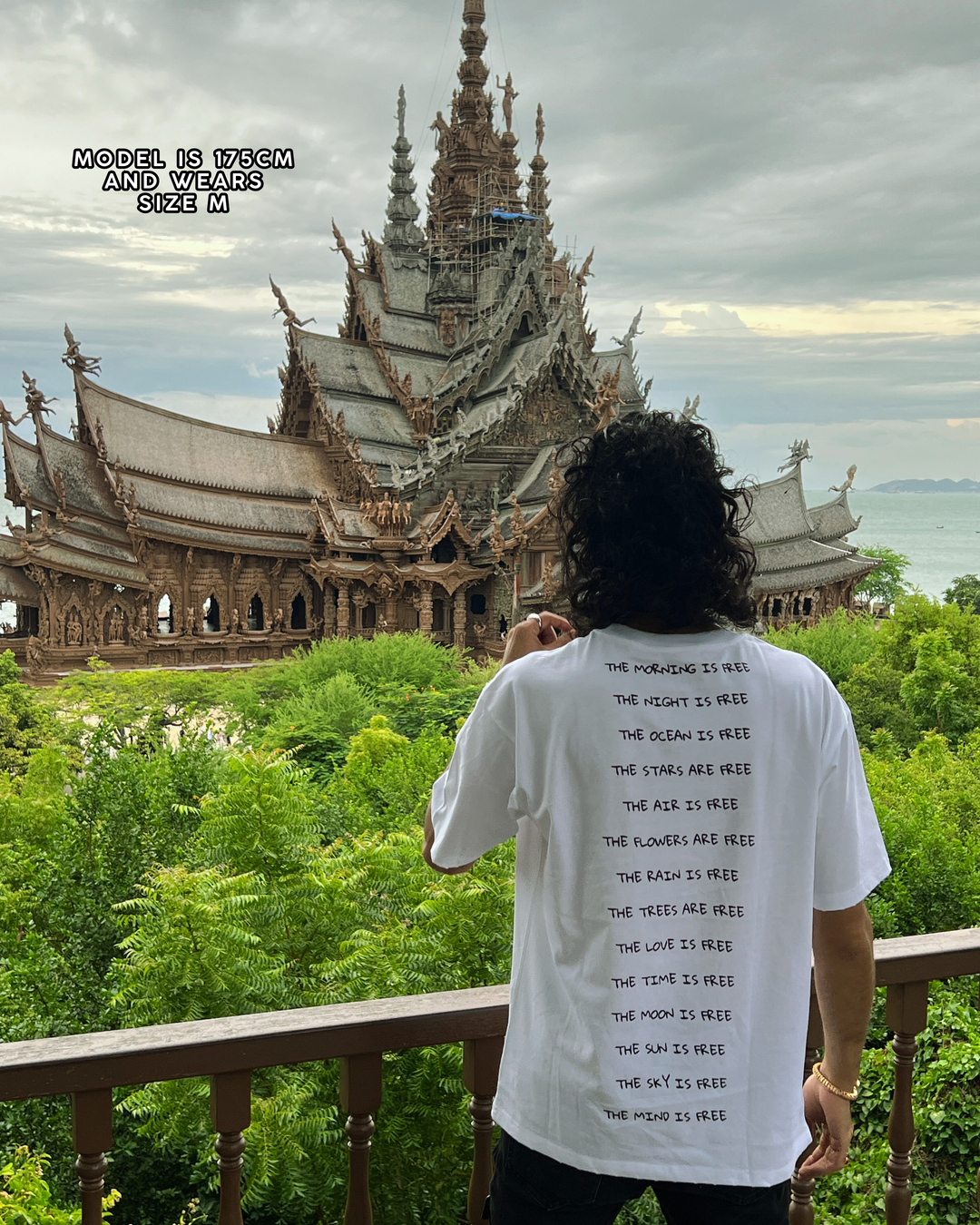 Person wearing a white t-shirt with text in front of a large, intricate temple surrounded by greenery.