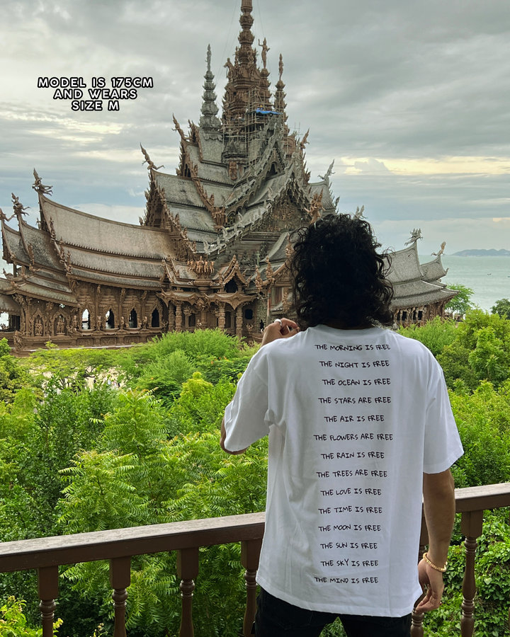 Person wearing a white t-shirt with text in front of a large, intricate temple surrounded by greenery.