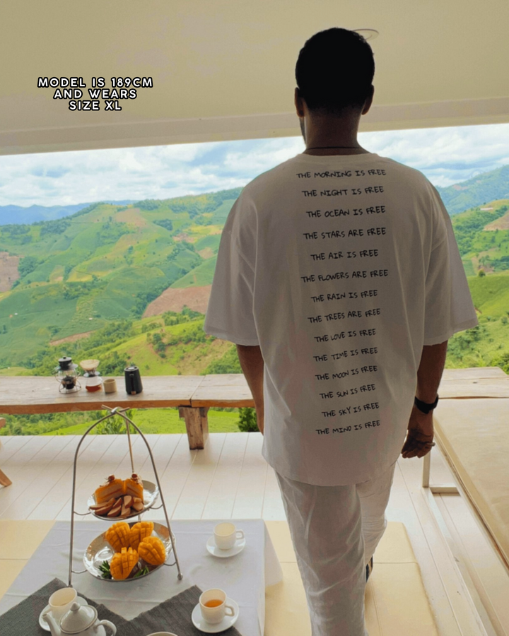 Person wearing a white t-shirt with text, standing in a room with a scenic view of mountains.