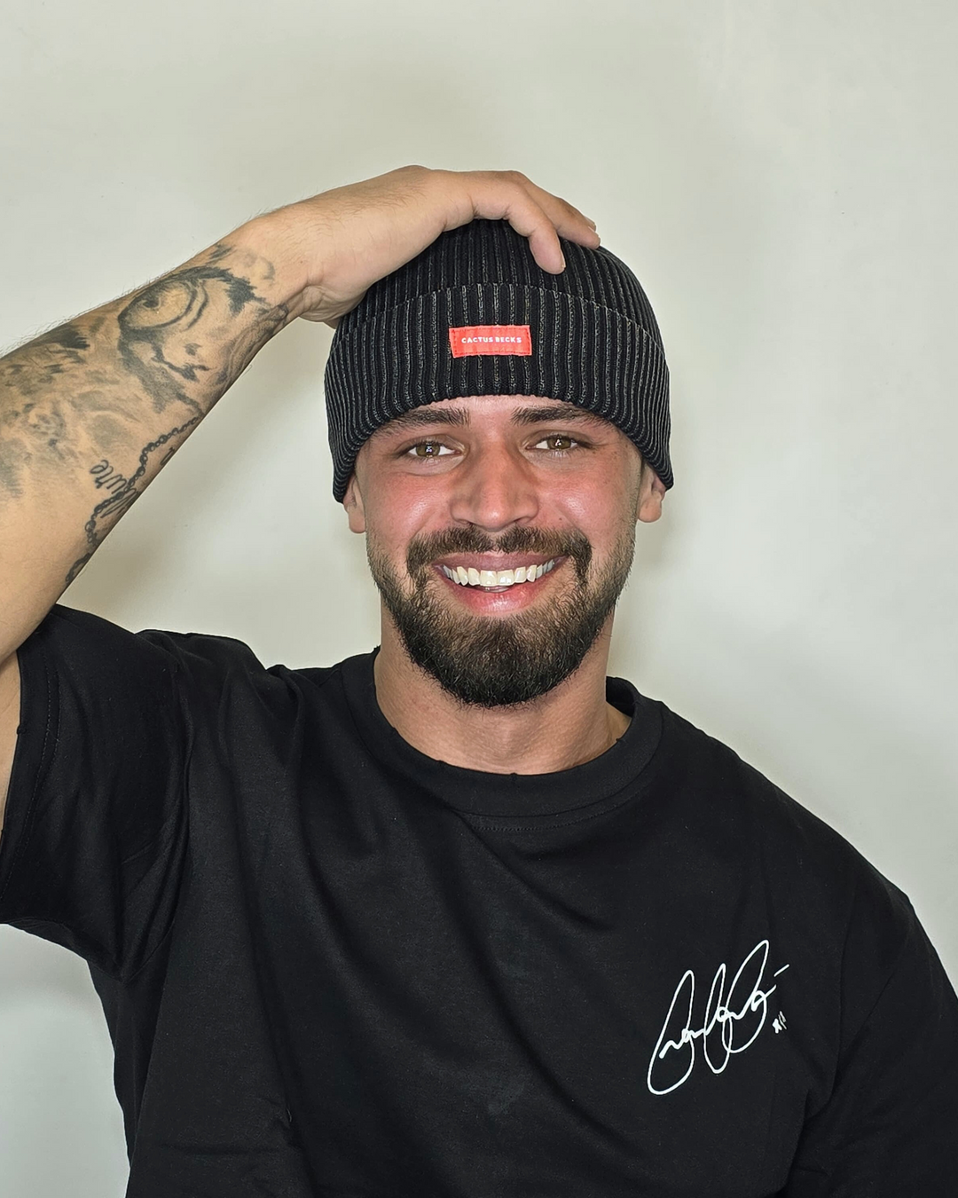 Man wearing a black beanie with a red logo and a black t-shirt with white text, against a plain background.