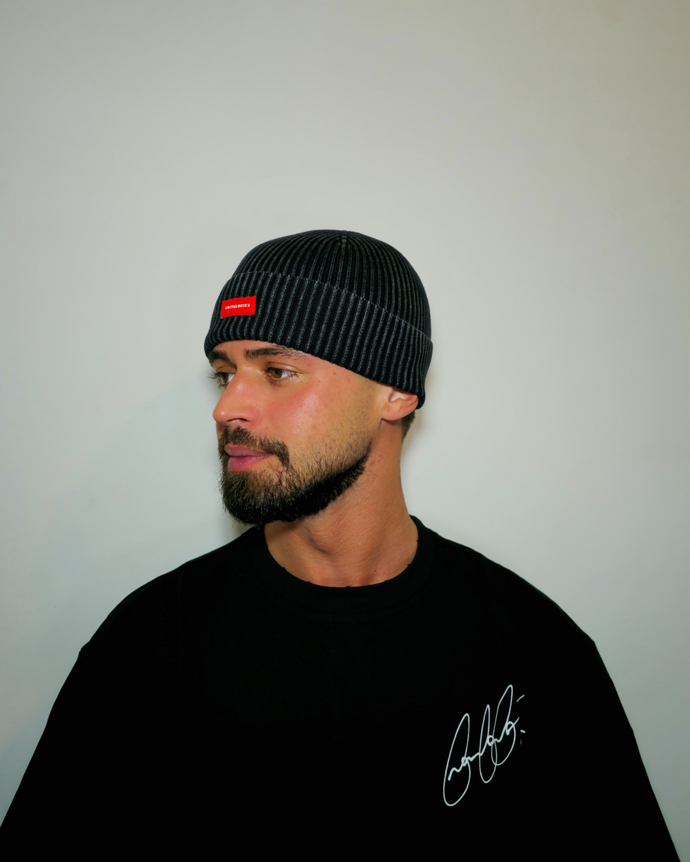 Man wearing a black beanie with a red logo and black shirt with signature, against a plain background